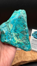Load image into Gallery viewer, A hand holds a large, rough turquoise-colored mineral specimen with green and blue marbling. A small product card labeled CHYSOX is visible on a wooden surface against a dark background.