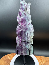 Load image into Gallery viewer, Hand-carved Rainbow Fluorite eagle with snake sculpture displayed on a custom stand, showcasing the multicolor green, purple, and blue banding of the natural stone.

