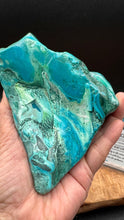 Load image into Gallery viewer, A hand holding a polished raw turquoise stone with swirling patterns of bright turquoise, teal, green, and white, set against a dark background.
